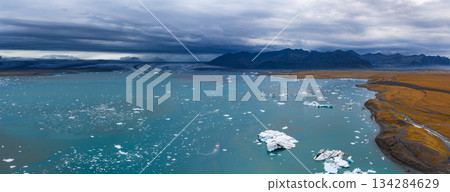 Turquoise waters of Jokulsarlon Glacier Lagoon with floating icebergs, a small boat, a glacier, rugged mountains, and golden brown terrain. Turquoise waters of Jokulsarlon Glacier Lagoon with floating icebergs, a small boat, a glacier, rugged mountains, and golden brown terrain. 134284629
