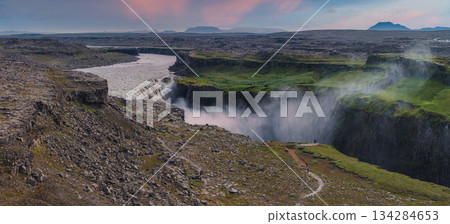 Dettifoss waterfall plunges into a deep canyon with mist rising, surrounded by rocky terrain, green moss, and a pink and blue sky in Iceland. Dettifoss waterfall plunges into a deep canyon with mist rising, surrounded by rocky terrain, green moss, and a pink and blue sky in Iceland. 134284653