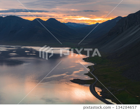 A winding road follows a reflective body of water at sunset, bordered by green fields and steep mountains under a vibrant orange and yellow sky. A winding road follows a reflective body of water at sunset, bordered by green fields and steep mountains under a vibrant orange and yellow sky. 134284657