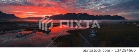 A serene harbor in Iceland at sunset, featuring boats, industrial buildings, calm water, and mist covered jagged mountains in the background. 134284660