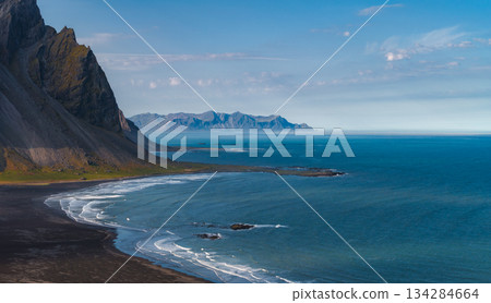The Stokknes Peninsula features black sand beaches, Vestrahorn mountain, turquoise ocean waters, and a clear blue sky in a dramatic Icelandic scene. 134284664