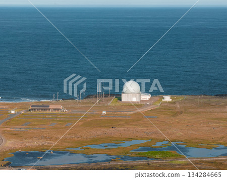 A white spherical radar dome near the ocean, surrounded by flat grassy terrain, wetlands, small water pools, and visible utility poles in Iceland. 134284665