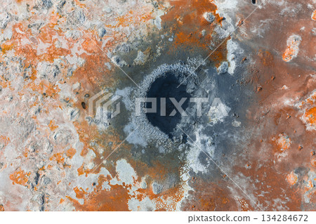 Aerial image of a bubbling geothermal mud pot in Iceland, surrounded by orange, brown, and white mineral deposits, showcasing unique geological textures. 134284672