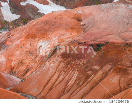 Multicolored rhyolite hills with orange, red, and brown hues, contrasted by green moss and white snow, showcasing erosion patterns and smooth slopes. 134284676
