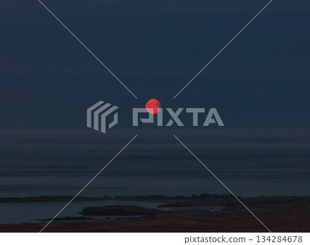 A vivid red moon rises over a calm, dark ocean at twilight, with faint landforms and rocky outcroppings visible along the Icelandic coastline. A vivid red moon rises over a calm, dark ocean at twilight, with faint landforms and rocky outcroppings visible along the Icelandic coastline. 134284678