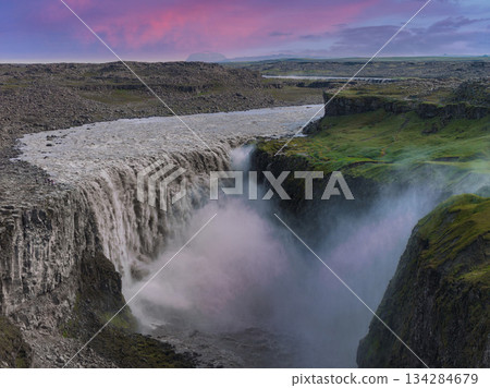 Dettifoss waterfall cascades into a deep canyon with rugged cliffs and green moss, set against a pink and purple sunset sky in Iceland. Dettifoss waterfall cascades into a deep canyon with rugged cliffs and green moss, set against a pink and purple sunset sky in Iceland. 134284679