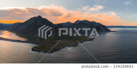 Aerial view of Iceland's Stokknes Peninsula with Vestrahorn's jagged peaks, a black sand beach, calm ocean waters, and a lighthouse at sunset. Aerial view of Iceland's Stokknes Peninsula with Vestrahorn's jagged peaks, a black sand beach, calm ocean waters, and a lighthouse at sunset. 134284691