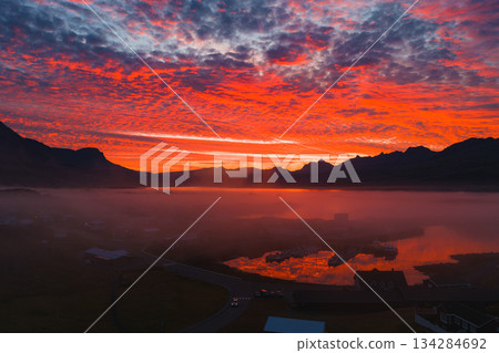 A vivid sunset illuminates the sky in red, orange, and purple over a misty Icelandic town. Still water reflects the scene, framed by jagged peaks. A vivid sunset illuminates the sky in red, orange, and purple over a misty Icelandic town. Still water reflects the scene, framed by jagged peaks. 134284692