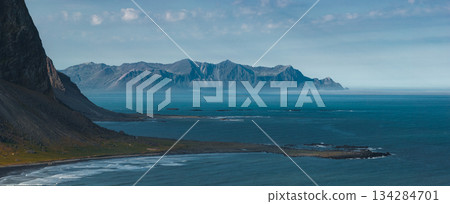 The image captures the Stokknes Peninsula in Iceland, featuring jagged mountains, a black sand beach, gentle waves, and a clear blue sky. 134284701
