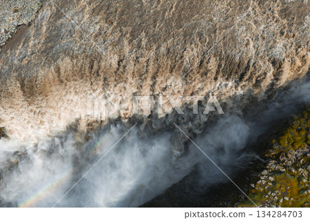 Dramatic aerial view of Dettifoss waterfall in Iceland, featuring turbulent muddy water, mist with a faint rainbow, and rocky terrain with mossy greenery. Dramatic aerial view of Dettifoss waterfall in Iceland, featuring turbulent muddy water, mist with a faint rainbow, and rocky terrain with mossy greenery. 134284703