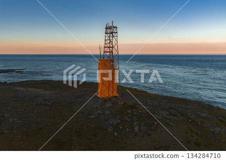 A vibrant orange lighthouse with a metal framework stands on a rocky coastal landscape. Calm ocean waters meet a sky of pink, orange, and blue hues. A vibrant orange lighthouse with a metal framework stands on a rocky coastal landscape. Calm ocean waters meet a sky of pink, orange, and blue hues. 134284710