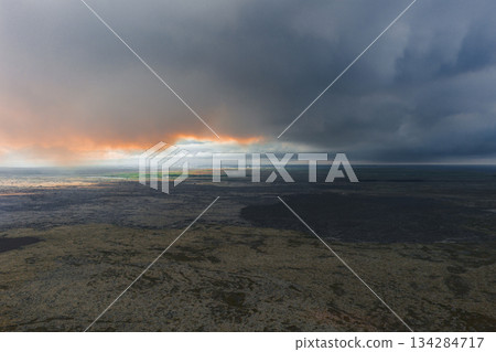 A dramatic aerial view of Iceland's volcanic terrain with dark rocks, mossy patches, and sunlight breaking through heavy clouds on the horizon. 134284717