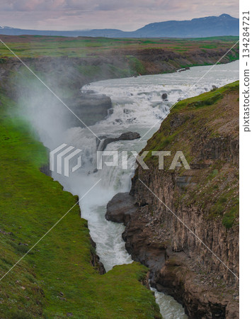 Gullfoss waterfall in Iceland flows through a rugged canyon, surrounded by green cliffs. Mist rises as distant mountains sit under a cloudy sky. 134284721