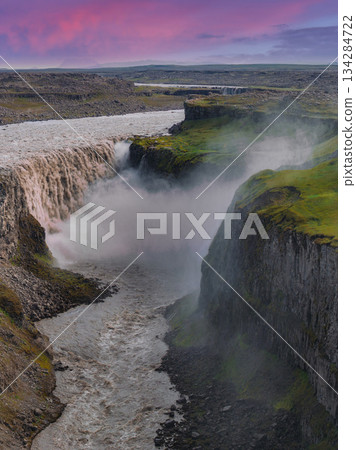 Dettifoss waterfall cascades into a deep canyon with moss covered cliffs, mist rising dramatically, under a pink and purple sky in Iceland. 134284722