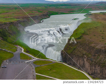 Gullfoss waterfall cascades in two tiers, surrounded by cliffs and greenery. Viewing platforms, pathways, and a parking area are visible in the scene. 134284723