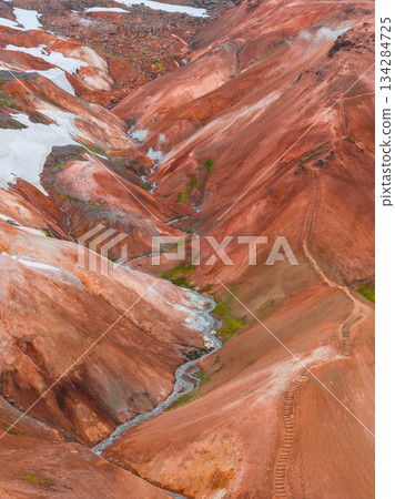 Vibrant red, orange, and brown rhyolite mountains in Landmannalaugar, Iceland, with snow patches, steam vents, a winding trail, and a small stream. 134284725