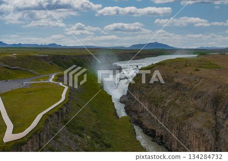 Gullfoss waterfall in Iceland flows into a rugged canyon with mist rising. A pathway and platform are visible, surrounded by green grass and cliffs. 134284732