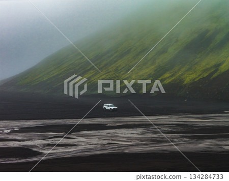 A white vehicle sits on black volcanic sand, with textured patterns in the foreground and a misty, moss covered hill in the background in Iceland. A white vehicle sits on black volcanic sand, with textured patterns in the foreground and a misty, moss covered hill in the background in Iceland. 134284733
