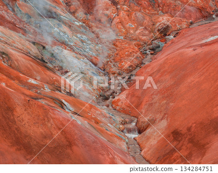 Geothermal terrain in Iceland with vivid red and orange hues, rising steam, and a small stream cutting through the mineral rich surface. 134284751