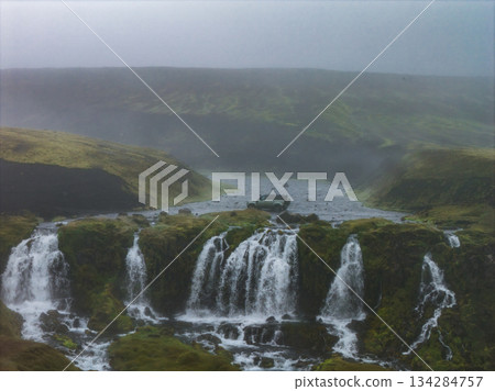 A cascading waterfall flows through moss covered terrain in Iceland. A vehicle is parked near a river under an overcast sky with mist and fog. A cascading waterfall flows through moss covered terrain in Iceland. A vehicle is parked near a river under an overcast sky with mist and fog. 134284757