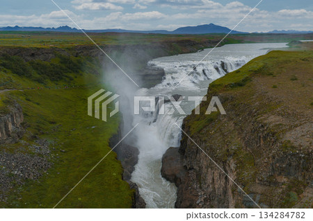Gullfoss waterfall in Iceland flows in two tiers into a canyon, surrounded by green terrain, mist, rolling hills, and distant mountains under a cloudy sky. 134284782
