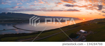 Aerial view of Akureyri, Iceland, during sunset. Features a calm fjord, curved bridge, shoreline town, green hills, and distant snow capped mountains. Aerial view of Akureyri, Iceland, during sunset. Features a calm fjord, curved bridge, shoreline town, green hills, and distant snow capped mountains. 134284788