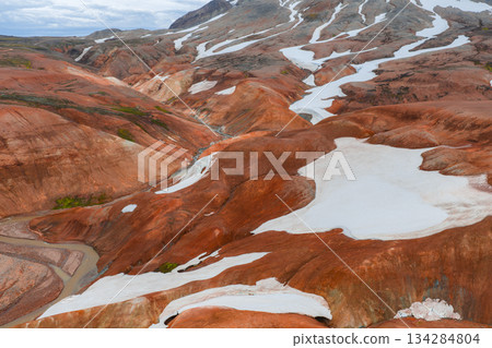 Vibrant red, orange, and brown rhyolite hills of Iceland's Rainbow Mountains with white snow patches and a winding stream cutting through the terrain. Vibrant red, orange, and brown rhyolite hills of Iceland's Rainbow Mountains with white snow patches and a winding stream cutting through the terrain. 134284804