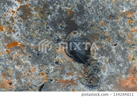 Aerial image of a dark oval shaped mud pot in Iceland, surrounded by cracked terrain with gray, orange, and rust hues, showcasing geothermal activity. 134284811