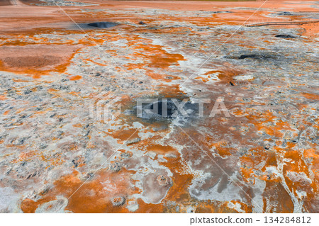 Aerial view of a geothermal area in Iceland featuring orange and white mineral deposits, steaming mud pots, fumaroles, and a barren, textured landscape. 134284812