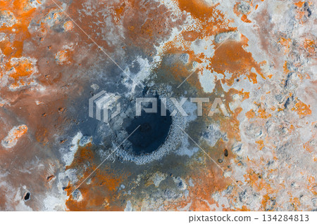 Aerial image of a geothermal area in Iceland featuring a dark crater like center surrounded by orange, white, and gray mineral deposits. 134284813