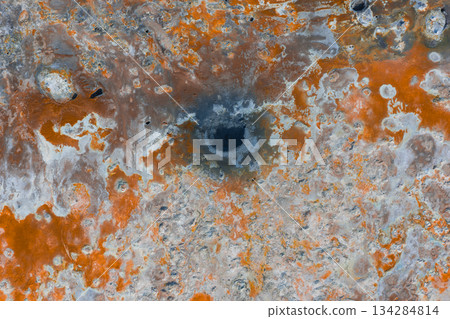 Aerial image of a geothermal area in Iceland featuring vibrant orange, white, and gray terrain, a dark mud pot, mineral deposits, and visible steam. 134284814