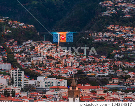 Aerial twilight view of Funchal, Madeira, terracotta rooftops on steep valleys, blue and gold viaduct panel with red cross, white houses, church towers, dusk light. Aerial twilight view of Funchal, Madeira, terracotta rooftops on steep valleys, blue and gold viaduct panel with red cross, white houses, church towers, dusk light. 134284817