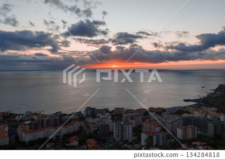 Funchal, Madeira, Portugal at dusk shows mid rise apartments with red roofs by the water, a rocky headland, and a small harbor as soft twilight light reflects on calm sea 134284818
