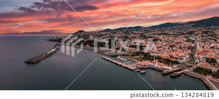 Aerial view shows Funchal, Madeira, Portugal at sunset, with breakwater and cruise pier framing marina, red tile rooftops on hills, calm water, and evening light. 134284819
