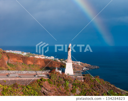 Cristo Rei at Garajau stands on a pedestal above the Atlantic in Madeira, Portugal. Terraced roads, coastal scrub, and white houses appear as a rainbow arcs under post storm light. 134284823