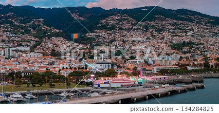 Aerial twilight view of Funchal, Madeira, with marina fairground, circus tent, rides, and an illuminated Portuguese coat of arms on the hillside, white houses on slopes. Aerial twilight view of Funchal, Madeira, with marina fairground, circus tent, rides, and an illuminated Portuguese coat of arms on the hillside, white houses on slopes. 134284825