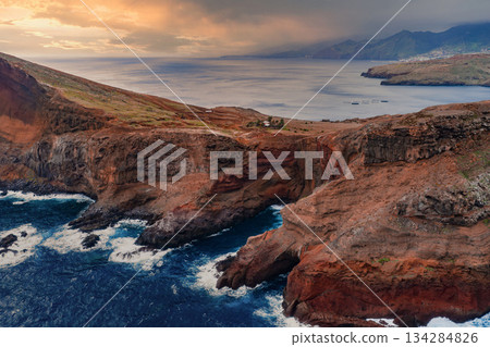 Aerial view shows rust colored cliffs and basalt headlands at Ponta de Sao Lourenco, Madeira, Portugal, waves crash, distant mountains, calm bay near Canical. Aerial view shows rust colored cliffs and basalt headlands at Ponta de Sao Lourenco, Madeira, Portugal, waves crash, distant mountains, calm bay near Canical. 134284826