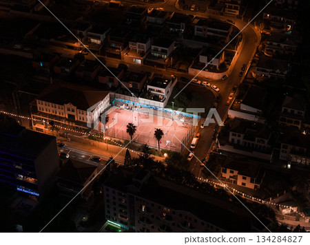 Aerial view shows a lit urban sports court in Funchal, Madeira, beside a white building, palm trees, parked cars, and warm streetlights along curving roads after dusk. 134284827