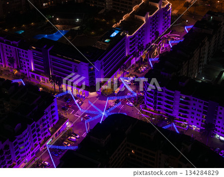 Aerial view of Funchal, Madeira at night, purple and blue lights zigzag above streets, mid rise apartments, parked cars, and a rooftop pool suggest a festive district 134284829