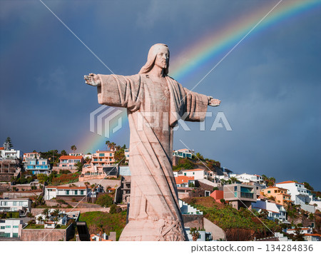 Cristo Rei statue stands on a hillside at Garajau, Madeira, Portugal, arms wide as a rainbow arcs behind. Dramatic cliffs and terraced homes descend toward the sea. 134284836