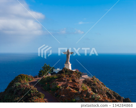 Cristo Rei statue at Garajau, Madeira, Portugal faces the Atlantic from a rocky promontory. Midday light, clear blue sea, winding path, terrace, and railing are visible. 134284868