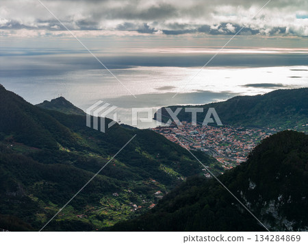 Aerial view of Madeira north coast near Machico or Santana, steep forested mountains meet a crescent bay, compact town by the water, soft late afternoon light. Aerial view of Madeira north coast near Machico or Santana, steep forested mountains meet a crescent bay, compact town by the water, soft late afternoon light. 134284869