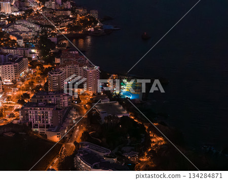 Aerial night scene in Funchal, Madeira, Portugal, shows hotel towers, curving orange streets, and a turquoise circular pool by the Atlantic, with reflections along the coast. Aerial night scene in Funchal, Madeira, Portugal, shows hotel towers, curving orange streets, and a turquoise circular pool by the Atlantic, with reflections along the coast. 134284871