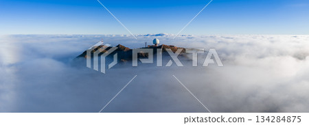 Aerial view shows Pico do Arieiro in Madeira, Portugal, with a white geodesic radar dome and antennas above a cloud layer, rocky ridge in clear daylight. 134284875