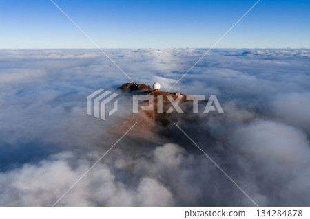 Aerial view shows Pico do Arieiro radar dome on Madeira Island, Portugal, above rolling clouds, with small buildings and paths visible in soft early or late light. 134284878