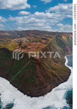 Aerial view of Madeira, Portugal, with sheer sea cliffs, waves at the base, fields and villages on the plateau, farm roads and stone walls, midday light and cumulus clouds. Aerial view of Madeira, Portugal, with sheer sea cliffs, waves at the base, fields and villages on the plateau, farm roads and stone walls, midday light and cumulus clouds. 134284881