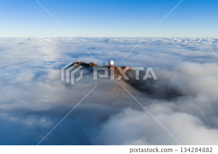 Aerial view shows Pico do Arieiro on Madeira Island with a white radar dome and ridge facilities above clouds at sunrise, soft light on rugged red brown rock and billowing clouds. Aerial view shows Pico do Arieiro on Madeira Island with a white radar dome and ridge facilities above clouds at sunrise, soft light on rugged red brown rock and billowing clouds. 134284882