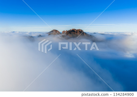 Rocky crest rises above a cloud layer at Pico do Arieiro, Madeira, aerial. Peaks appear to float in soft morning or late afternoon light with mist easing details. 134284900