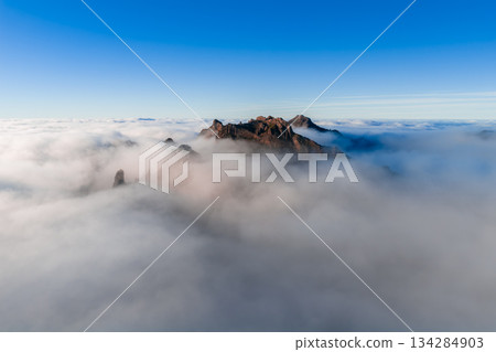 Aerial view shows Pico do Arieiro ridge slicing cloud inversion in Madeira, Portugal. Warm sunrise or sunset light bathes jagged peaks, rocky outcrops, and volcanic forms. Aerial view shows Pico do Arieiro ridge slicing cloud inversion in Madeira, Portugal. Warm sunrise or sunset light bathes jagged peaks, rocky outcrops, and volcanic forms. 134284903