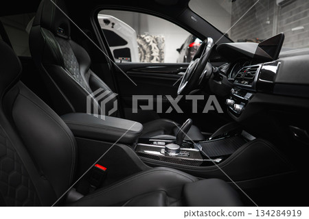 Black leather seats with quilted pattern, high tech dashboard, and carbon fiber accents in a modern car. Background shows a car shop setting. Black leather seats with quilted pattern, high tech dashboard, and carbon fiber accents in a modern car. Background shows a car shop setting. 134284919